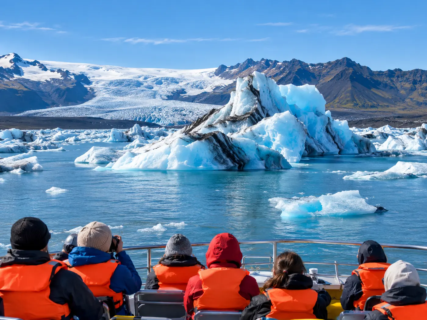 Passengers in orange life jackets seen from behind on the deck of an amphibian boat beside an iceberg