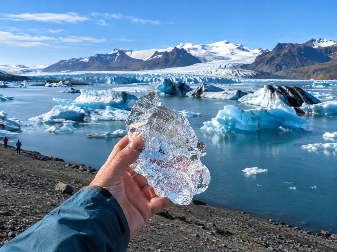 A hand holding a piece of clear glacier ice over the blue water of the lagoon