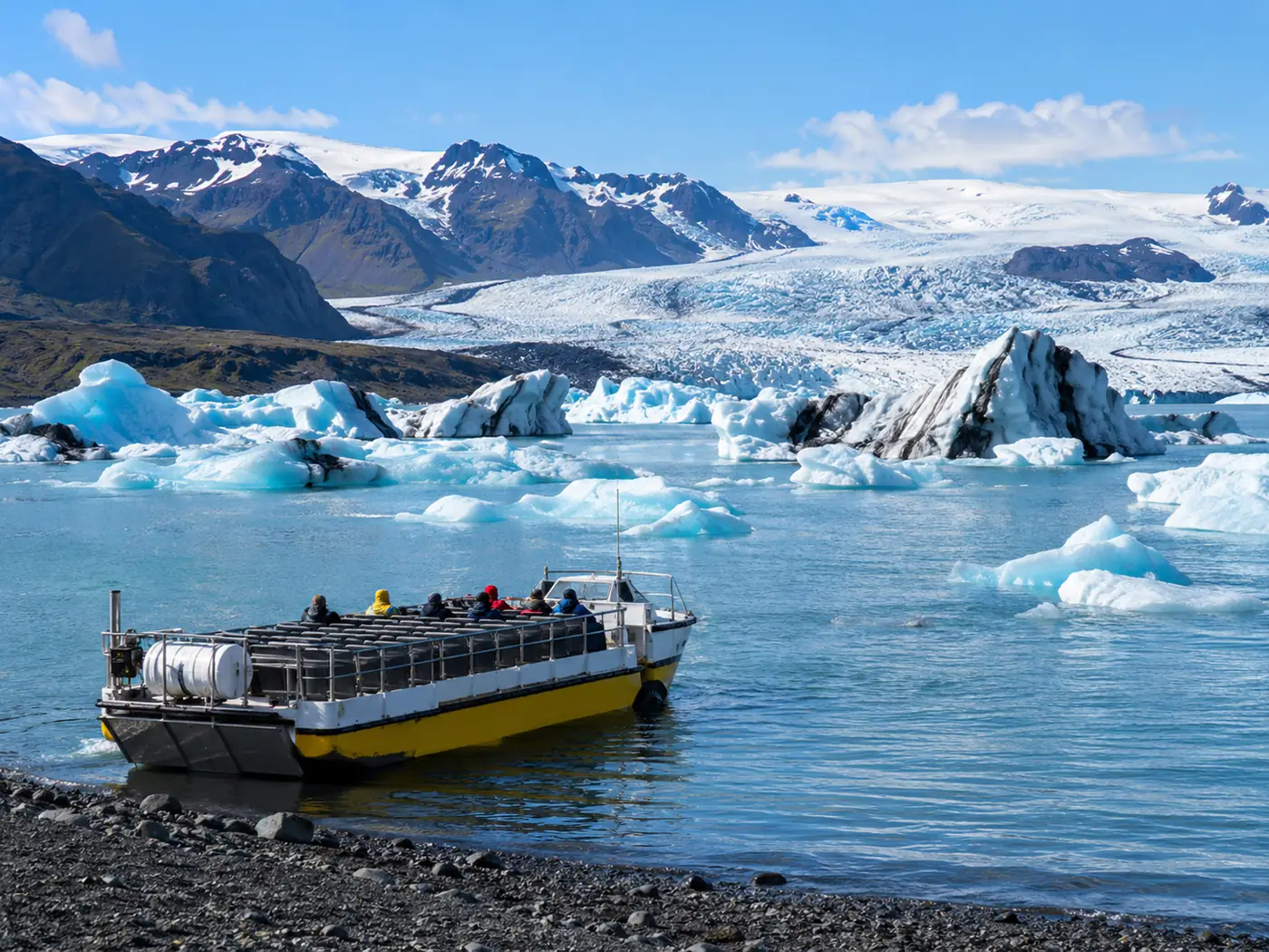 A wide amphibian boat on the lagoon seen from the shore, icebergs behind