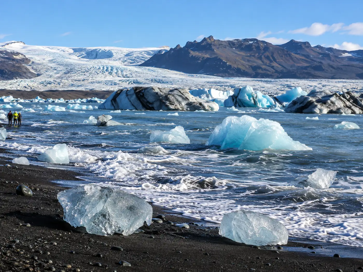 Surf washing around ice blocks on a black beach, seen from well back up the sand